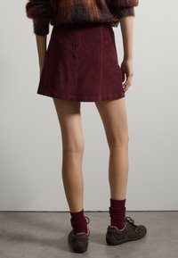 Gonna mini in corduroy bordeaux, con texture a costine, abbinata a scarpe scure e calzini bordeaux. Lo sfondo è minimale con una tonalità grigia.