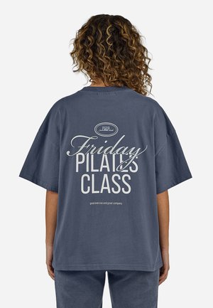 Osoba o kręconych włosach ubrana w luźny ciemnoszary T-shirt z napisem "Friday Pilates Class" na plecach, stojąca na jednolitym tle.