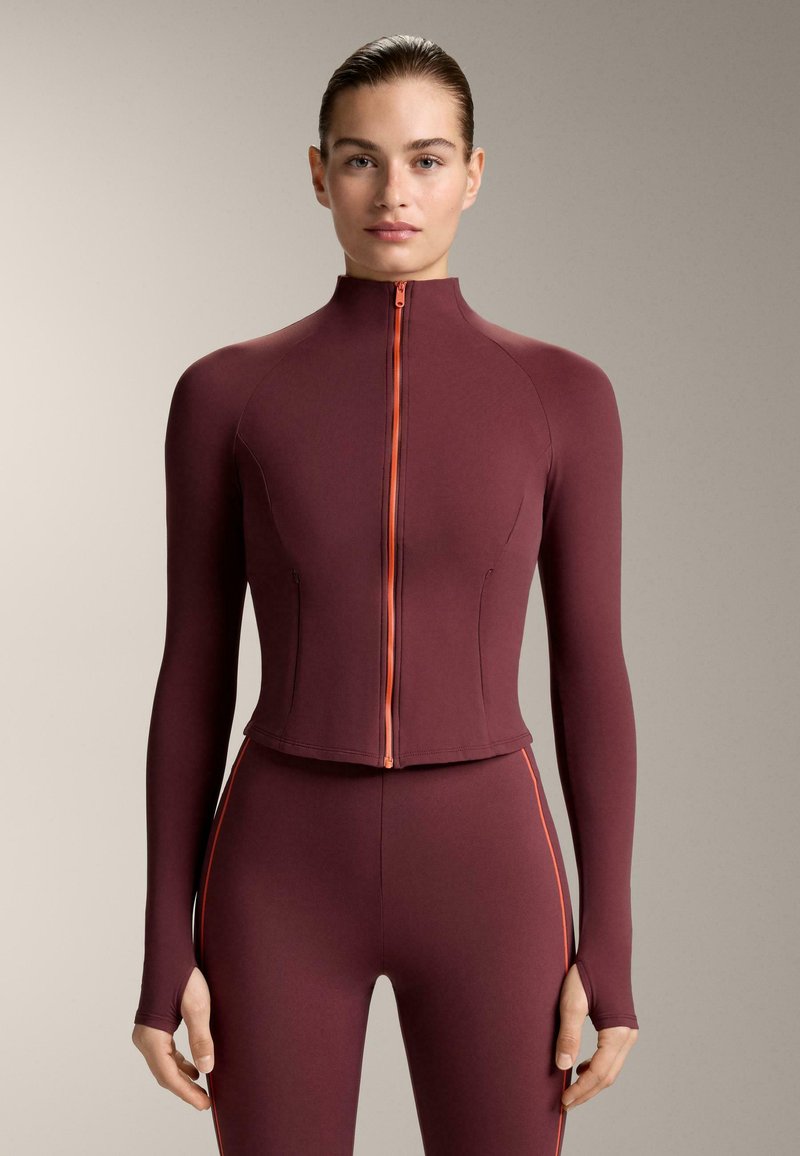 Fahéjvörös testhez simuló sportfelső magas nyakkal és narancssárga cipzárral, hosszú ujjal, ujjlyukkal, hozzáillő testhez simuló nadrággal.