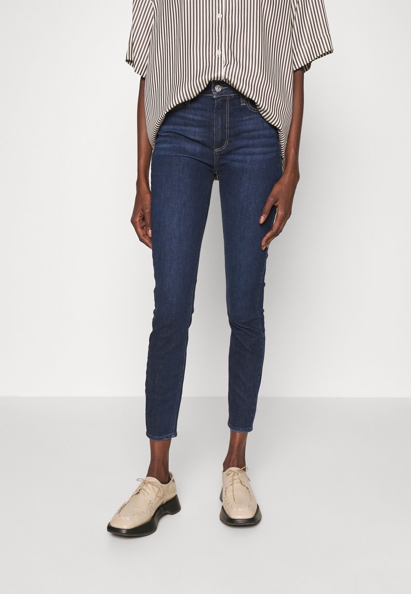 Mörkblå skinny jeans med en slät textur och subtila sömnadsdetaljer, tillsammans med ljusbeige snörskor. Ränderad skjorta ovanpå.