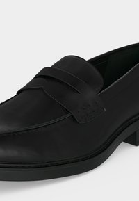 Mocassin en cuir noir avec un petit talon, une couture de style mocassin et une bride décorative sur le dessus du pied, sur fond blanc.