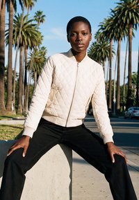 Giovane con capelli corti che indossa una giacca imbottita bianca e pantaloni neri, seduto su un blocco di cemento con palme sullo sfondo.