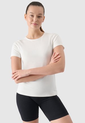 Joven mujer de pie con los brazos cruzados, vistiendo una camisa blanca de manga corta y pantalones cortos de ciclismo negros sobre un fondo claro y liso.