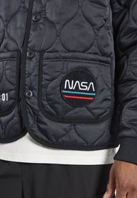 Chaqueta acolchada negra con cierre de botones, que presenta un parche de NASA con bandas de color y tejido texturizado. Incluye dos bolsillos frontales.