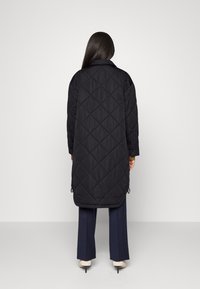 Žena s dlouhými tmavými vlasy stojí zády a nosí dlouhý černý prošívaný kabát, námořnické kalhoty a bílé lodičky na vysokém podpatku na jednobarevném pozadí.