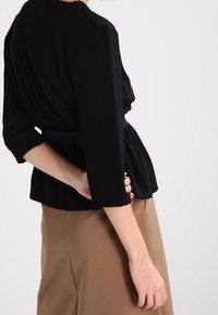 Zwarte, getailleerde top met driekwartmouwen, verstelbare taille en zachte textuur, gecombineerd met een beige rok. Eenvoudig design zonder patronen.