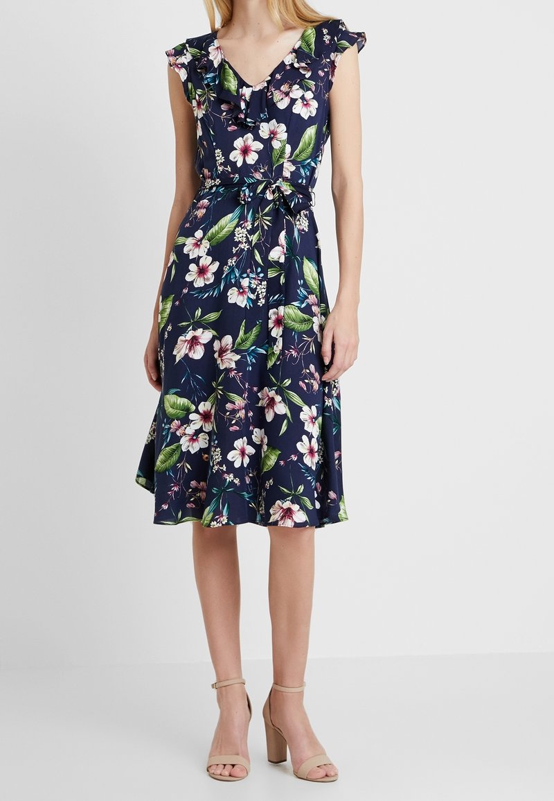 Robe fleurie bleu marine avec manches courtes volantées, encolure en V et taille ceinturée. Présente des fleurs blanches et des feuilles vertes sur un tissu doux et fluide.