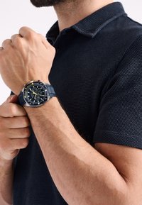 Hronogrāfs ar tumši zilu silikona siksniņu, apaļu nerūsējošā tērauda korpusu, melnu ciparnīcu, dzelteniem akcentiem un trim subciparnīcām.