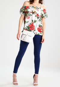 Kukkainen olkaimeton paita, jossa on punaisia ja keltaisia kukkia valkoisella taustalla, yhdistettynä tummansinisiin kapeutuisiin farkkuihin ja valkoiseen olkalaukkuun.