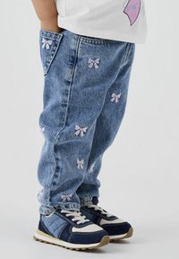 Hellblaue Jeans mit kleinen, rosa Schleifen-Stickereien, kombiniert mit marineblauen und weißen Sneakers mit strukturierten Sohlen.