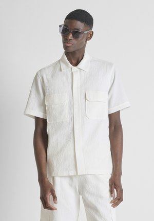 Jeune homme portant une chemise blanche texturée à manches courtes et un short assorti avec des lunettes de soleil sombres, debout devant un fond clair uni.