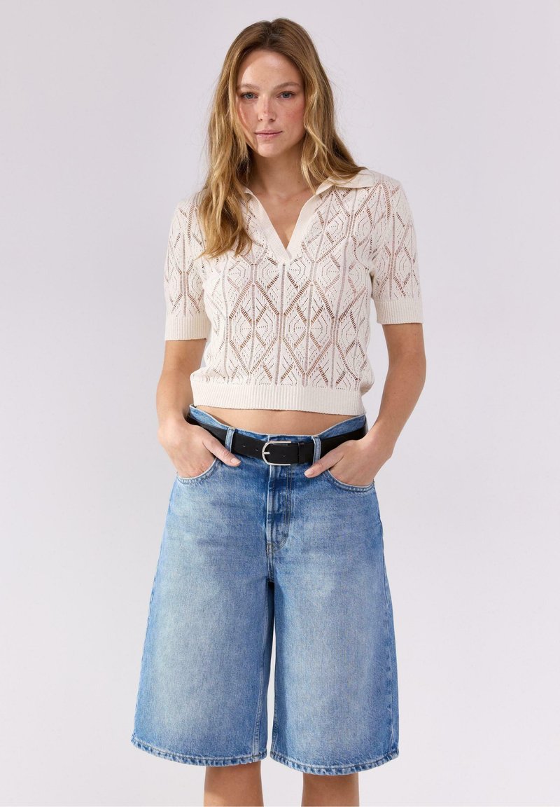 Maglione lavorato a maglia a maniche corte di colore crema con un motivo geometrico a lattice. Abbinato a pantaloni corti di jeans azzurri e una cintura nera.