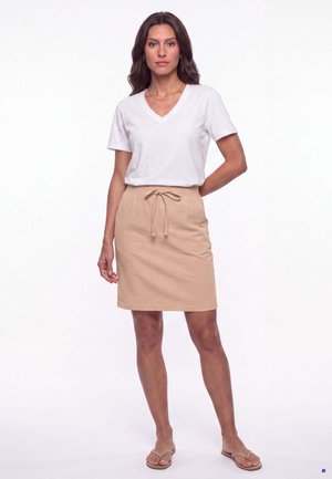 Kvinde står iført en hvid kortærmet V-hals T-shirt, beige knælange nederdel med snøre og beige sandaler mod en ensfarvet hvid baggrund.