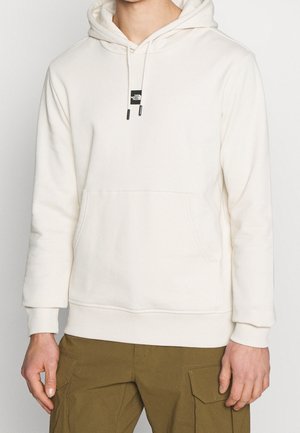 Persoon die een crèmekleurige hoodie draagt met een voorzak en het The North Face-logo, gecombineerd met olijfgroene cargobroeken.
