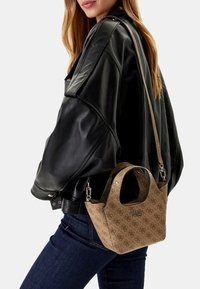 Donna con giacca di pelle nera e jeans scuri porta una borsa Guess a fantasia beige con manici corti e lunghi a spalla.