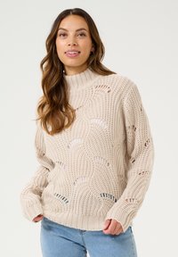 Pull en maille crème avec un col haut, présentant des motifs ajourés et des poignets côtelés, associé à un jean bleu clair.