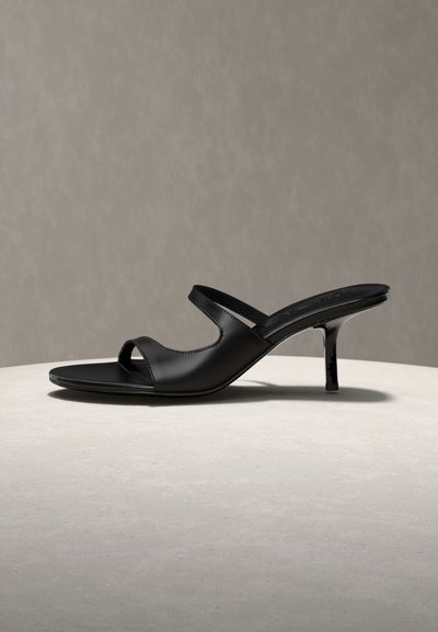 Mule noire en cuir avec talon, deux brides ouvertes, bout pointu, et talon stiletto fin de hauteur moyenne sur une surface claire et un fond neutre.