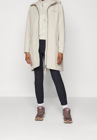 Manteau imperméable beige clair avec capuche, fermeture éclair intégrale et poches latérales. Porté par-dessus un haut blanc et associé à un pantalon foncé et des chaussures de randonnée marron.