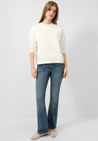 Witte sweatshirt met geribde manchetten en zoom, gecombineerd met lichtblauwe flare jeans. Model staat voor een neutrale achtergrond, met lichte schoenen aan.