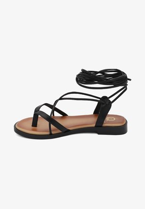 Sandalo nero con fasce sottili, dotato di suola piatta e di una morbida insolla beige. Lacci neri lunghi per legare alla caviglia.