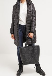 Osoba oblečená v černém prošívaném kabátu, bílém pólovém tričku, tmavých džínách a černých bootách, držící černou plátěnou tašku s embosovanými geometrickými vzory.