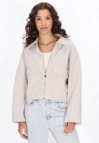 Giacca beige tagliata con cerniera frontale, ampie tasche a pattina e colletto. Tessuto liscio e vestibilità comoda, abbinata a jeans blu chiaro.