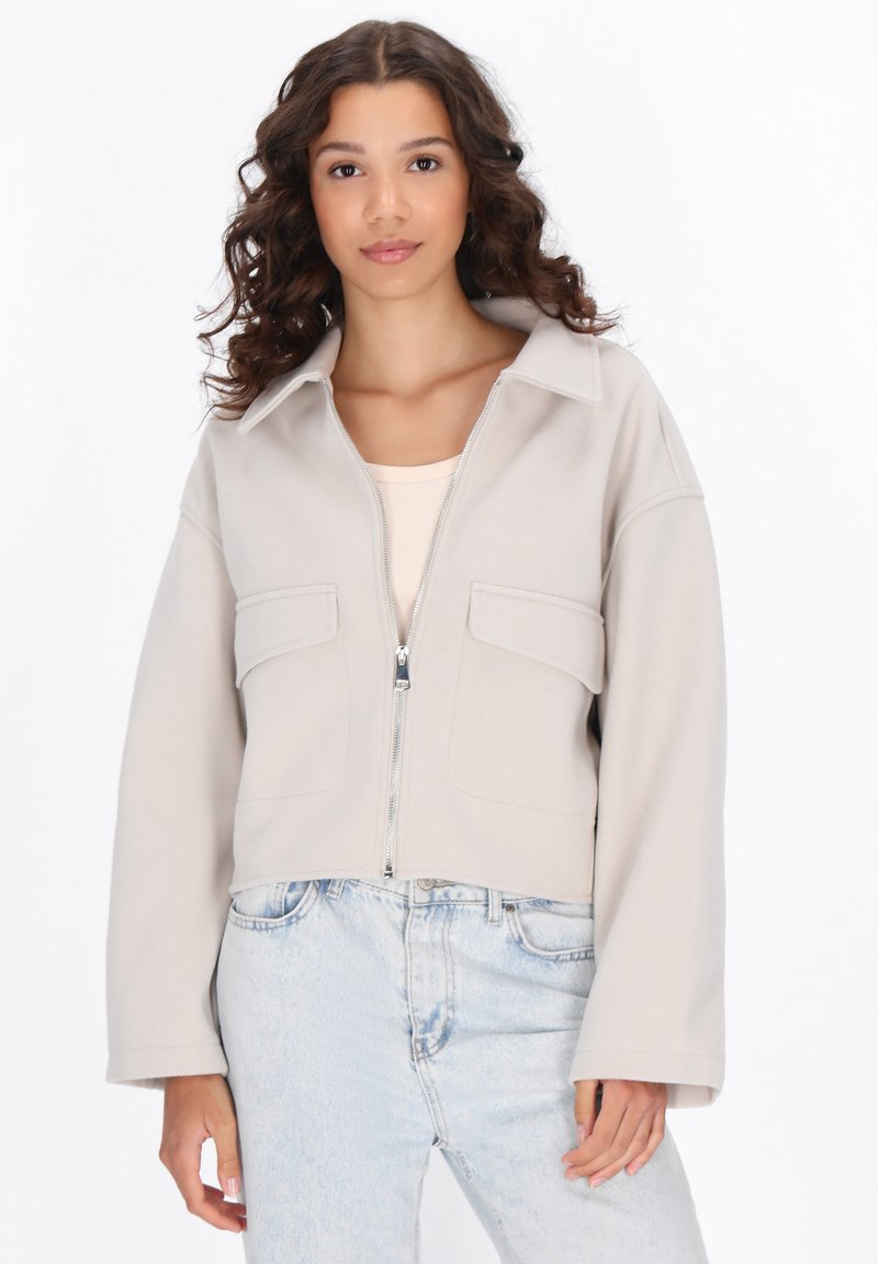 Giacca beige tagliata con cerniera frontale, ampie tasche a pattina e colletto. Tessuto liscio e vestibilità comoda, abbinata a jeans blu chiaro.
