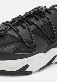 Czarne skórzane sneakersy z teksturowaną górną częścią, wyposażone w elastyczne sznurowadła i masywną białą podeszwę środkową. Projekt zawiera subtelne przeszycia i czarne akcenty.