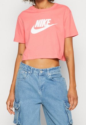 Personne portant un t-shirt Nike rose cropped et un pantalon cargo en denim bleu ample avec des poches, montrant le ventre et deux tatouages sur les bras.