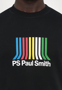 Chemise noire avec des rayures verticales multicolores en bleu, rouge, jaune, blanc et vert au-dessus du texte "PS Paul Smith" sur la poitrine.