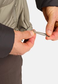 Veste extérieure grise avec un cordon de serrage à l'ourlet, ajustée par une main. Le tissu texturé et les coutures détaillées sont visibles.