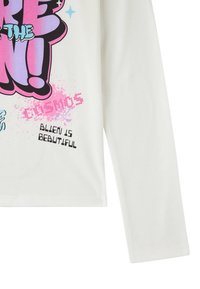 Maglietta bianca a maniche lunghe con grafica colorata che recita: "SEI FUORI DA QUESTO COSMO" in rosa, nero e blu, con dettagli di spruzzi giocosi.