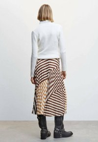 Un pull en maille blanche avec un col haut associé à une jupe midi plissée ornée d'un motif géométrique en rayures marron et beige. Bottes noires jusqu'aux genoux.