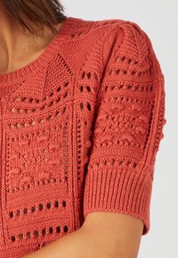 Maglione lavorato a maglia in corallo con motivi testurizzati, maniche corte e polsini a costine. Presenta dettagli con punto aperto per una migliore traspirabilità.