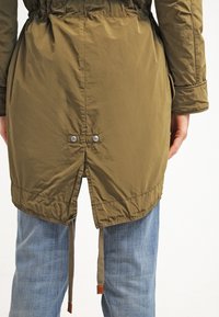 Primer plano de la parte posterior de una chaqueta verde oliva con una pequeña abertura y dos broches metálicos, usada sobre jeans azules.