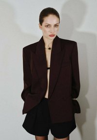 Blazer oversize color burdeos con solapas afiladas, un solo botón y bolsillos, combinado con pantalones cortos negros. Tejido suave y corte relajado.