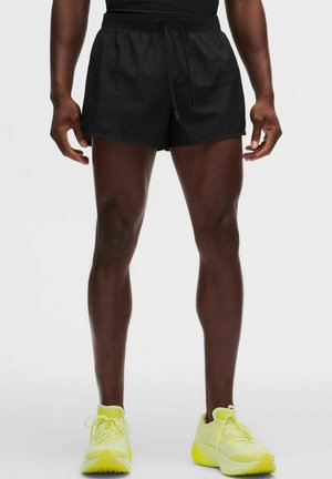 Mandlig atlet iført sorte løbeshorts og klare gule løbesko, stående mod en ensfarvet lys baggrund.