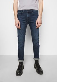 Tmavě modré džínové kalhoty slim fit se zapínáním na knoflík a zip, pěti kapsami a ohrnutými nohavicemi, doplněné černými šněrovacími botami.