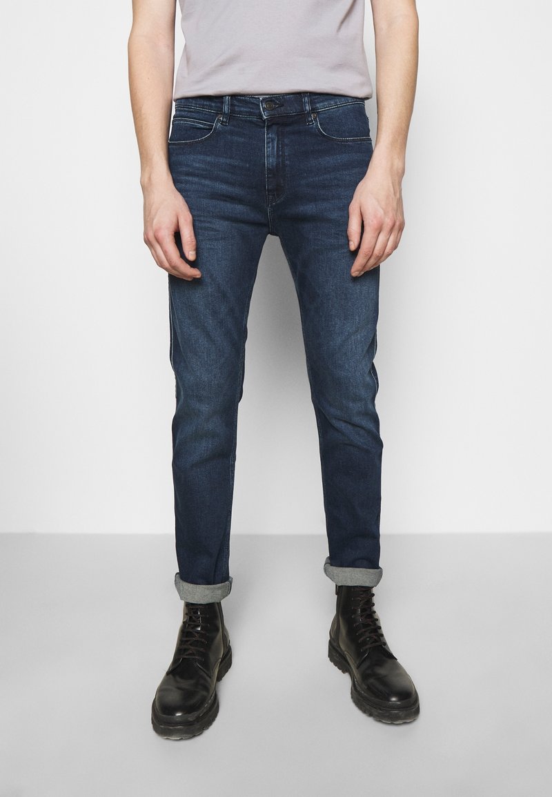 Tmavě modré džínové kalhoty slim fit se zapínáním na knoflík a zip, pěti kapsami a ohrnutými nohavicemi, doplněné černými šněrovacími botami.