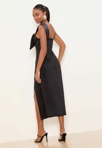 Robe midi noire avec un corsage ajusté, des bretelles larges en satin, une fente latérale et une texture lisse, associée à des sandales noires à talons hauts.