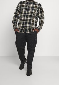 Rutig flanellskjorta i marinblått, svart och gult med knappstängning. Matchas med svarta cargobyxor med sidofickor och avslappnad passform.