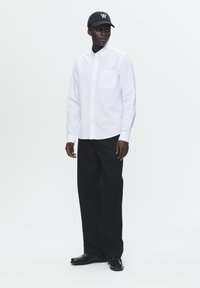 Camisa blanca de botones con un bolsillo en el pecho, combinada con pantalones negros de pierna ancha y zapatos negros. Una gorra negra con un logo blanco completa el atuendo.