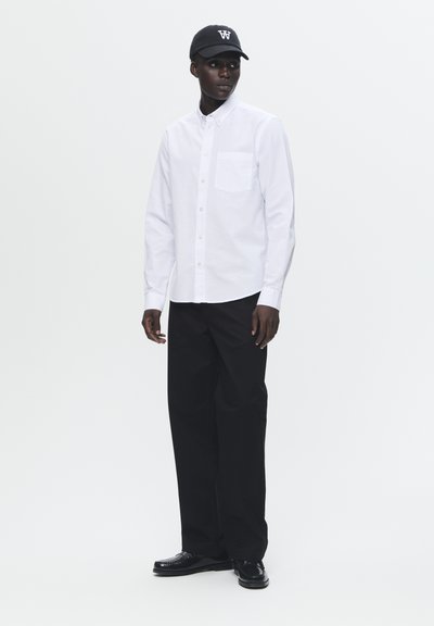 Hvid skjorte med knapper og en brystlomme, parret med sorte vide bukser og sorte sko. En sort kasket med et hvidt logo fuldender outfittet.