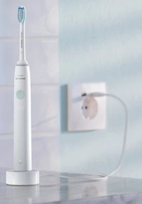 Spazzolino elettrico bianco con testina blu, base tonda e pulsante di accensione verde, collegato a una presa di corrente in un ambiente dai colori chiari.