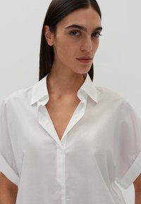 Camicia bianca over-size in tessuto leggero, con colletto a punta e maniche corte arrotolate, dotata di una chiusura frontale a bottoni.