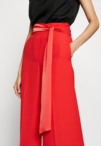 Červené kalhoty s širokými nohavicemi a odpovídajícím opaskem. Vyrobeny z hladkého materiálu, s bočními kapsami a upraveným střihem pro strukturovaný vzhled.