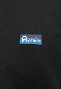 Μαύρο ύφασμα με μια ραμμένη ορθογώνια ετικέτα της μάρκας Penfield που παρουσιάζει ένα γραφικό ορεινού όγκου σε κόκκινο και μπλε χρώμα.