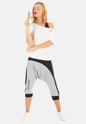 Weißes Off-Shoulder-Oberteil, graue und schwarze Haremshosen mit einem geschwungenen Design und schwarzen Akzenten. Das Model hält eine pinke Blume und trägt weiße Sneakers.