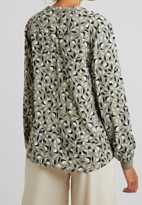 Femme portant une blouse à manches longues vert clair avec un motif de feuilles abstrait et un pantalon beige, vue de dos devant un fond uni.
