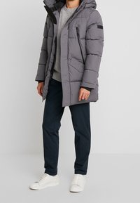 Piumino grigio con cappuccio, design trapuntato, chiusura frontale con zip, tasca sul petto con zip e tasche laterali. Indossato sopra un maglione grigio.
